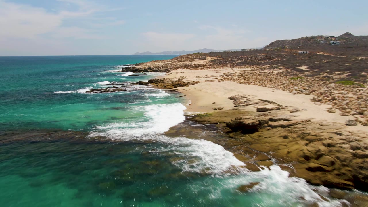 Aerial frame at Buzzards Beach with a wider sweep of white surf crossing dark rock shelves, clear turquoise shallows in front, dry sand to the right, and no visible people.