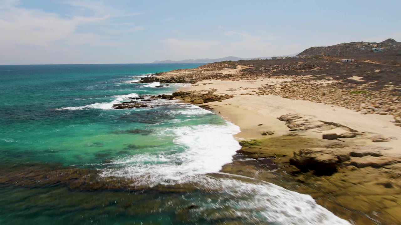 Aerial frame of Buzzards Beach showing a curved band of foam around a rocky point, green-blue water in the foreground, tan sand beyond the rocks, and no visible people.