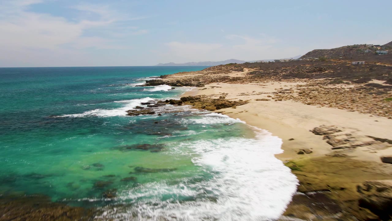 Aerial frame from Buzzards Beach with the coastline running diagonally, white water expanding over nearshore rocks, open sand along the right edge, and no visible people.