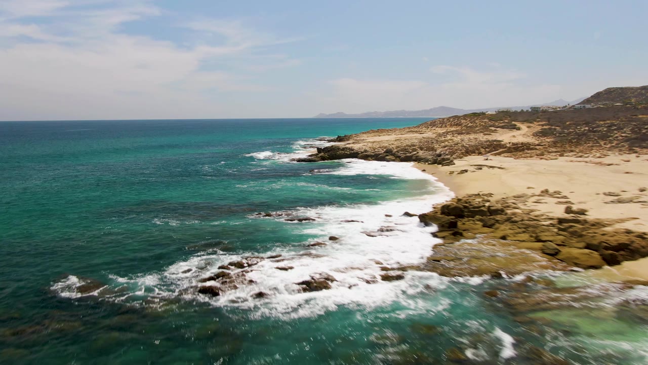 The aerial view at Buzzards Beach shows white surf spreading farther across dark shoreline rocks while clear blue-green water fills the foreground and no people are visible.
