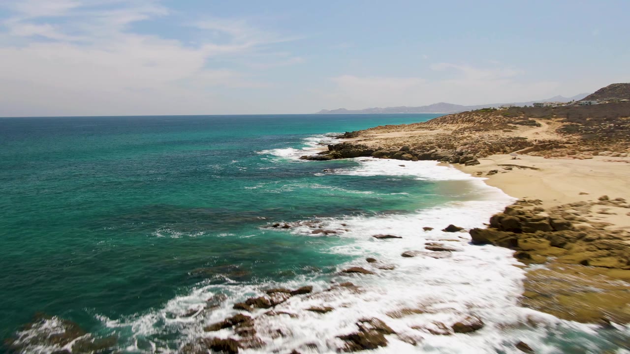 At 0:02, waves curl around the rocky point at Buzzards Beach, with a crescent of sand to the right, open ocean to the left, and no visible people.