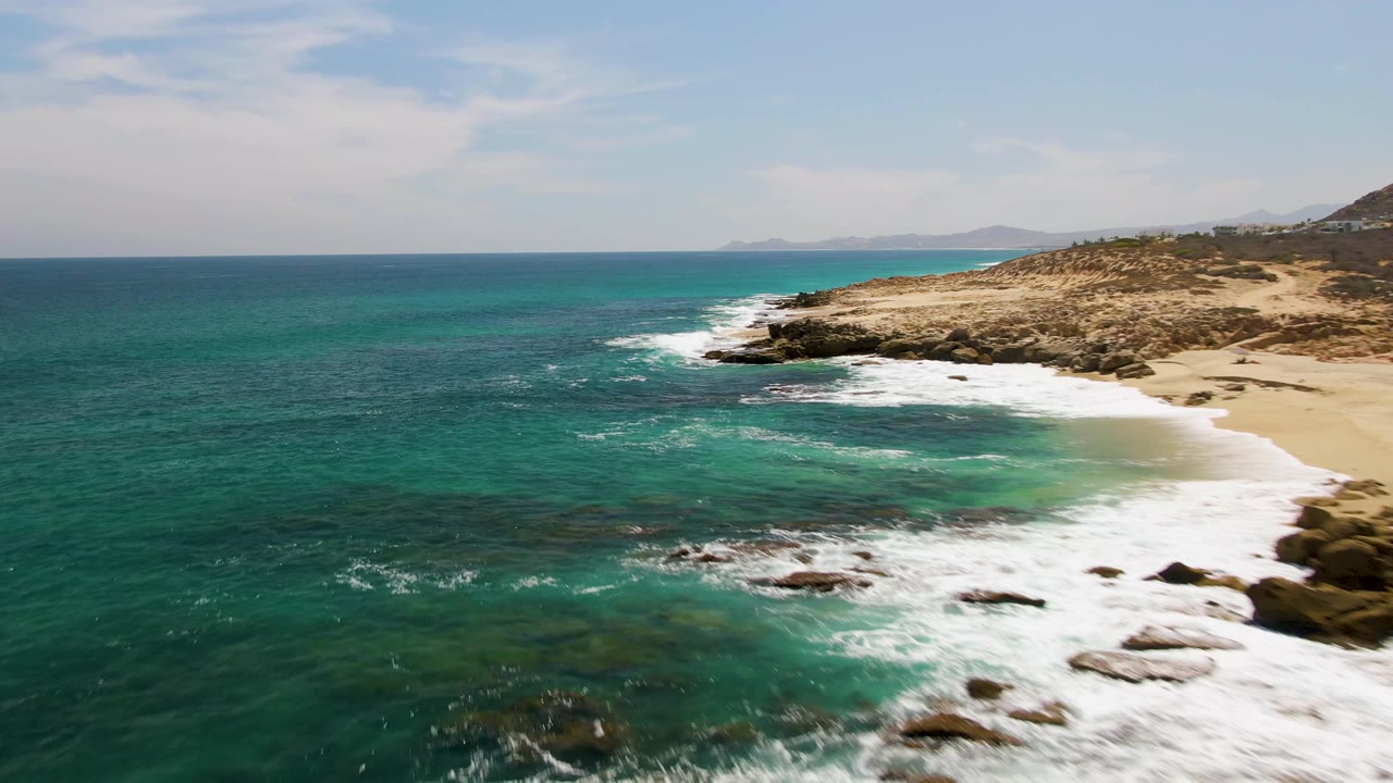 The Buzzards Beach shoreline is seen from above with turquoise shallows near the sand, white foam sliding along the beach edge, rocky outcrops, and no visible people.