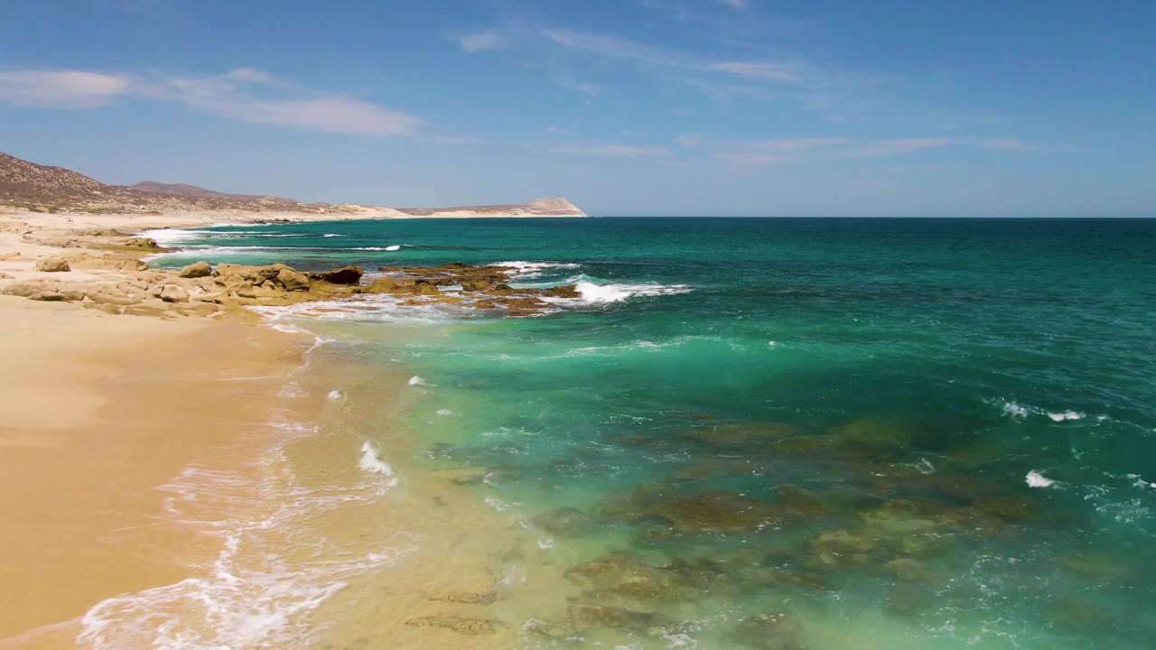 Turquoise waves reach a sandy and rocky shoreline under blue sky.