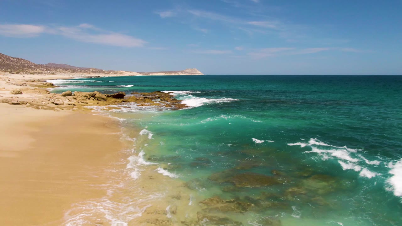 Buzzards Beach shoreline with a curved line of white foam advancing over tan sand, shallow turquoise water, exposed rocks, distant hills, and no visible people.