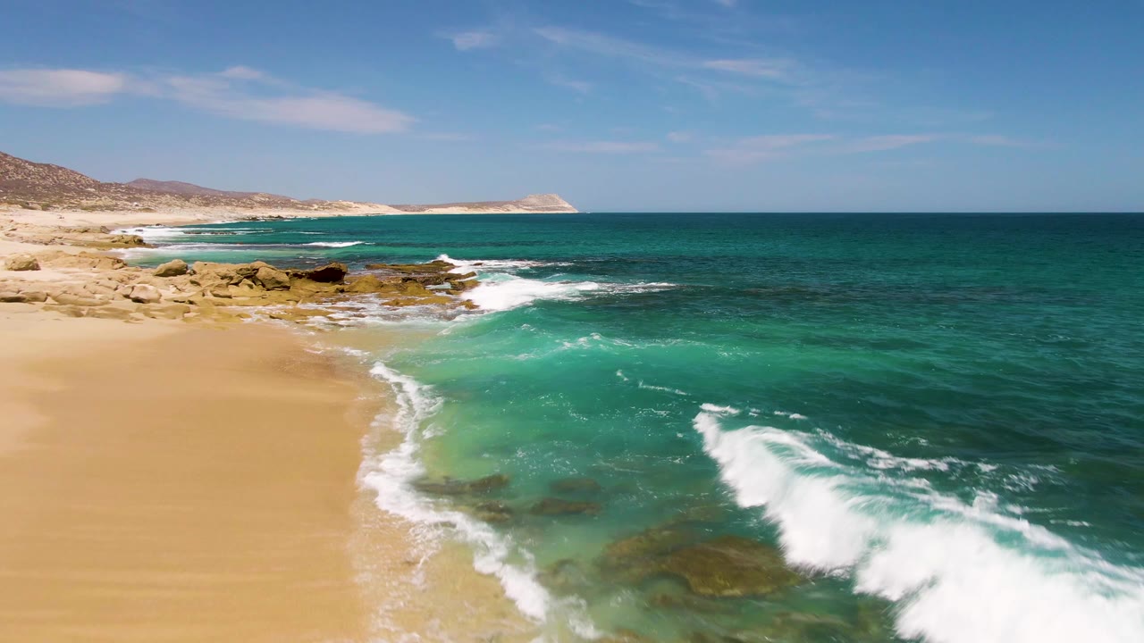 A stronger wave breaks on the right side of the Buzzards Beach frame while clear turquoise shallows, tan beach, rocky patches, distant dry hills, and no visible people remain in view.