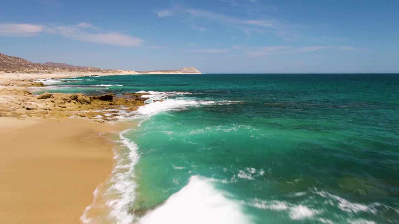 Mid-clip frame at Buzzards Beach with white foam spreading across the lower shoreline, green-blue water filling the cove, tan sand to the left, rocks ahead, and no visible people.