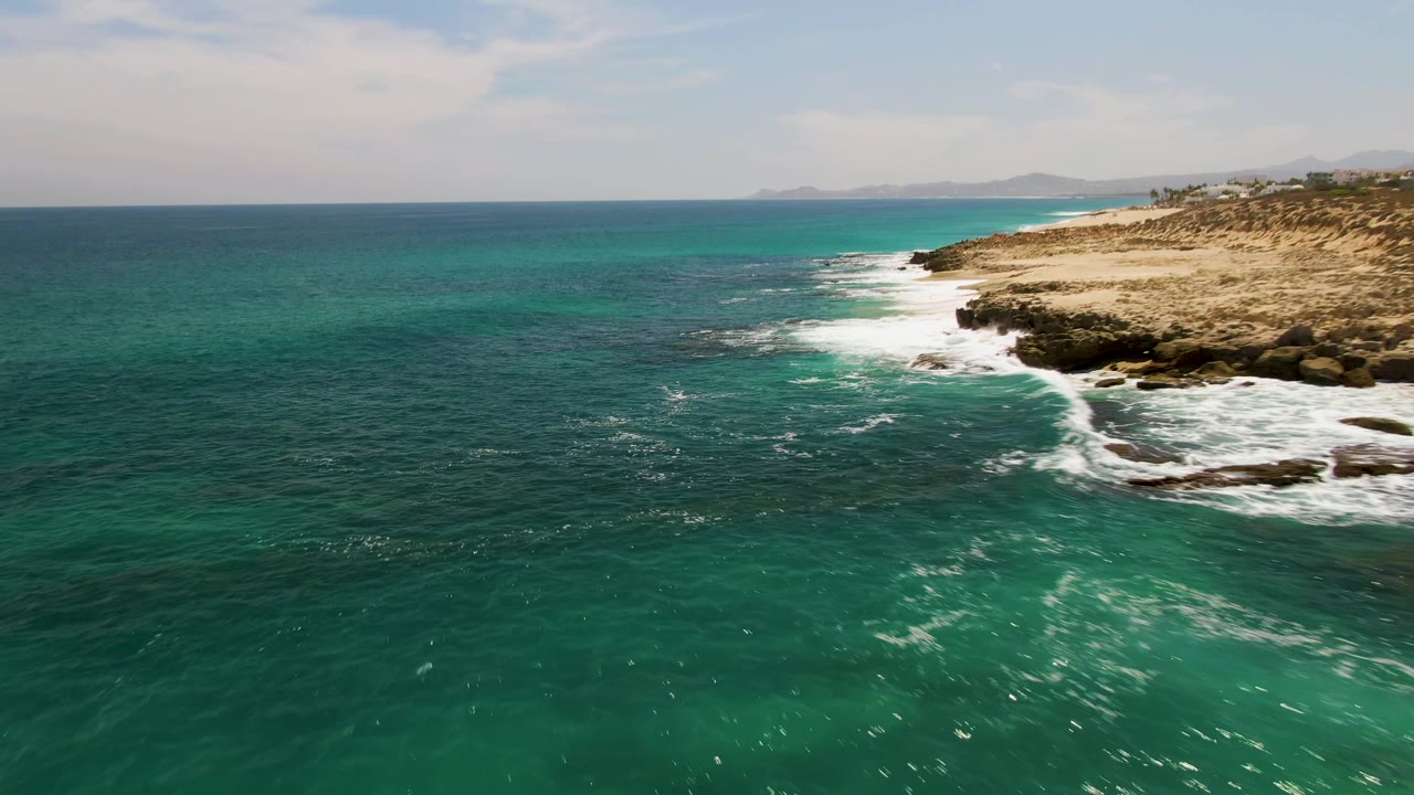 Aerial view at Buzzards Beach with green-blue ocean in the foreground, a band of white foam hitting black rocks, tan coastal ground to the right, and no visible people.
