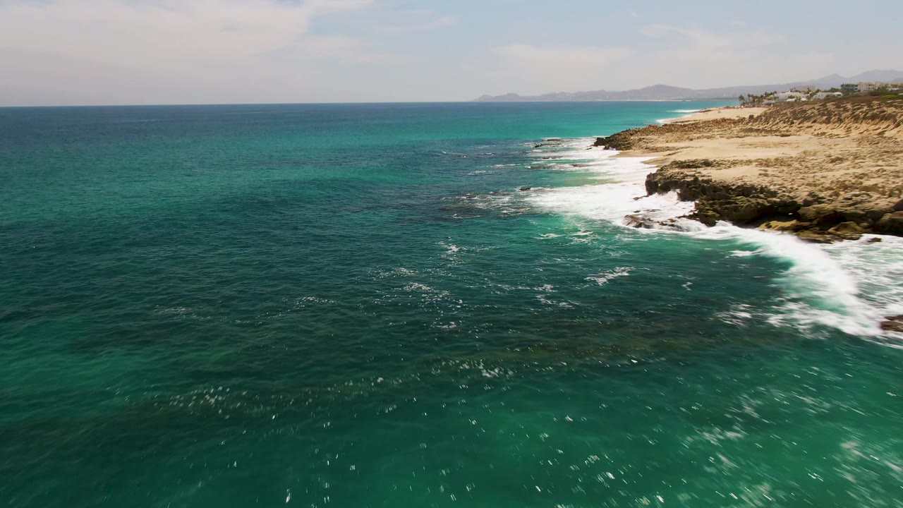 Wide drone frame from Buzzards Beach showing a long diagonal shoreline, turquoise water over darker reef patches, white breakers at the rocks, distant hazy hills, and no visible people.