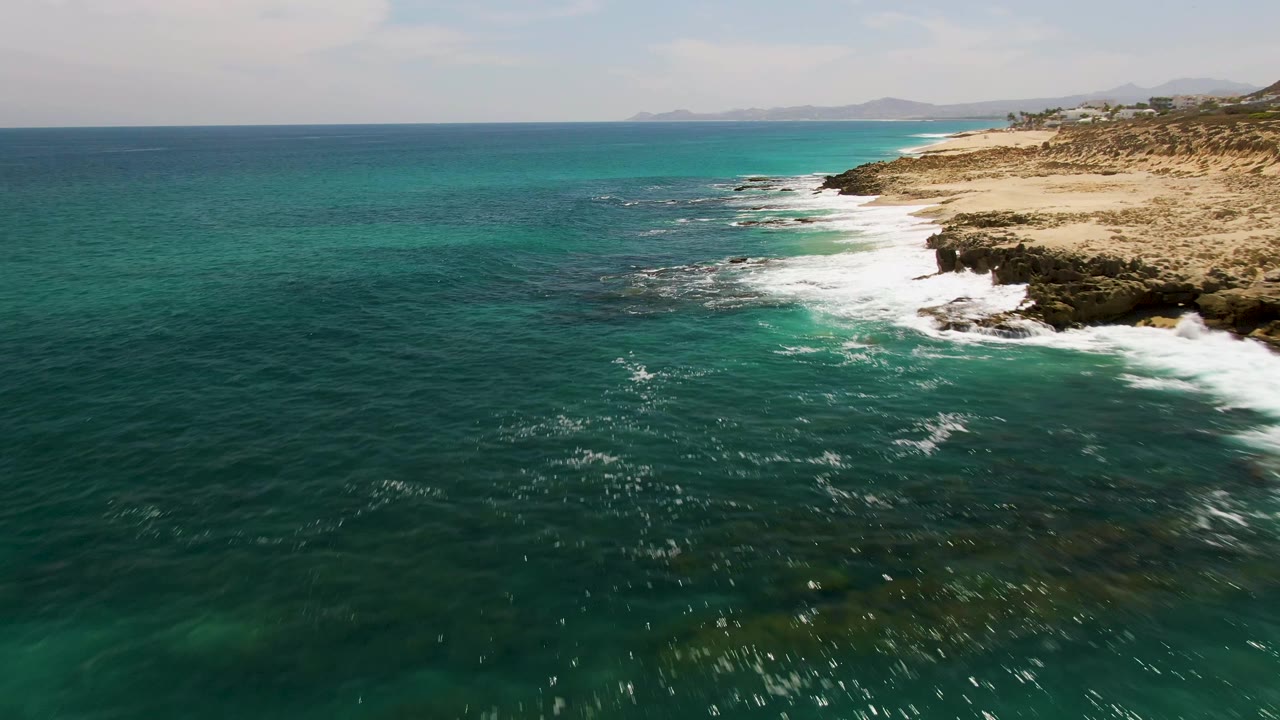 Aerial frame over Buzzards Beach with open teal water in the foreground, white foam spreading along the rocky edge, tan land on the right, and no visible people.