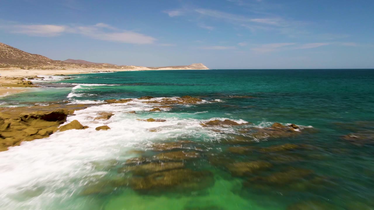 Aerial view of waves breaking over rocky turquoise shoreline beside a sandy beach.