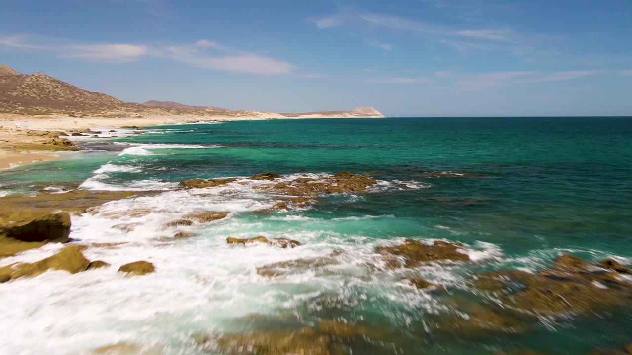 At Buzzards Beach in Baja California Sur, foamy waves cover more of the foreground rocks while the sandy beach and dry hills remain visible to the left, with no visible people.