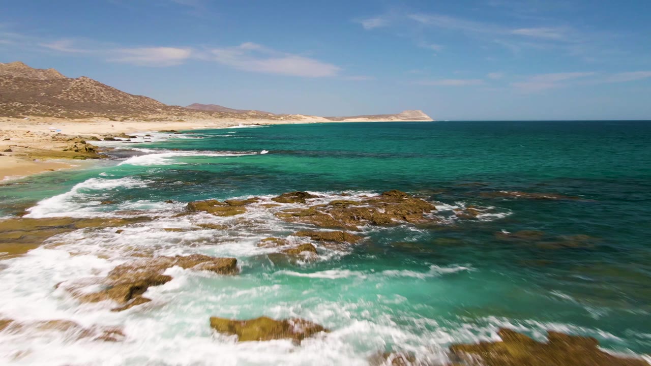 Buzzards Beach coastline appears from an aerial angle as surf breaks over low rock shelves, with green-blue shallows, sand, hills, open sea, and no visible people.