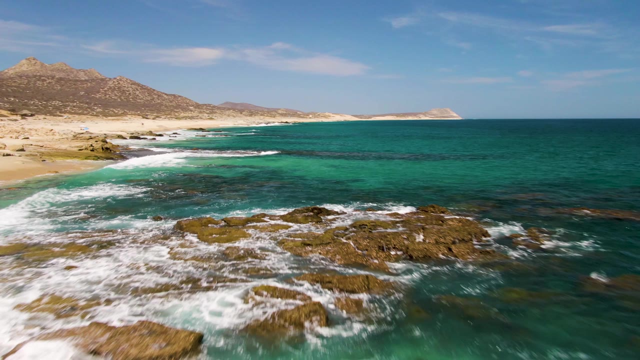 Aerial frame from Buzzards Beach in Baja California Sur showing foreground rocks ringed by white foam, clear turquoise water, sandy coastline, hills, and no visible people.