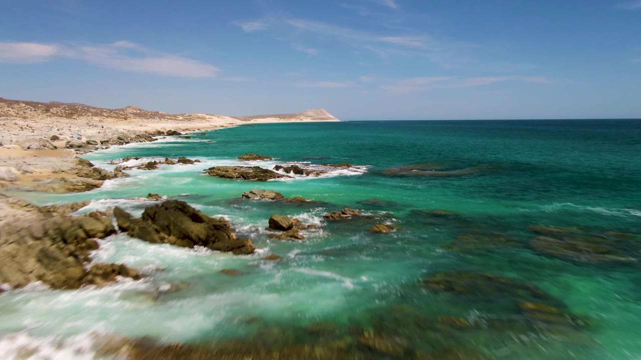 Turquoise surf breaking around rocky outcrops beside a sandy coast.
