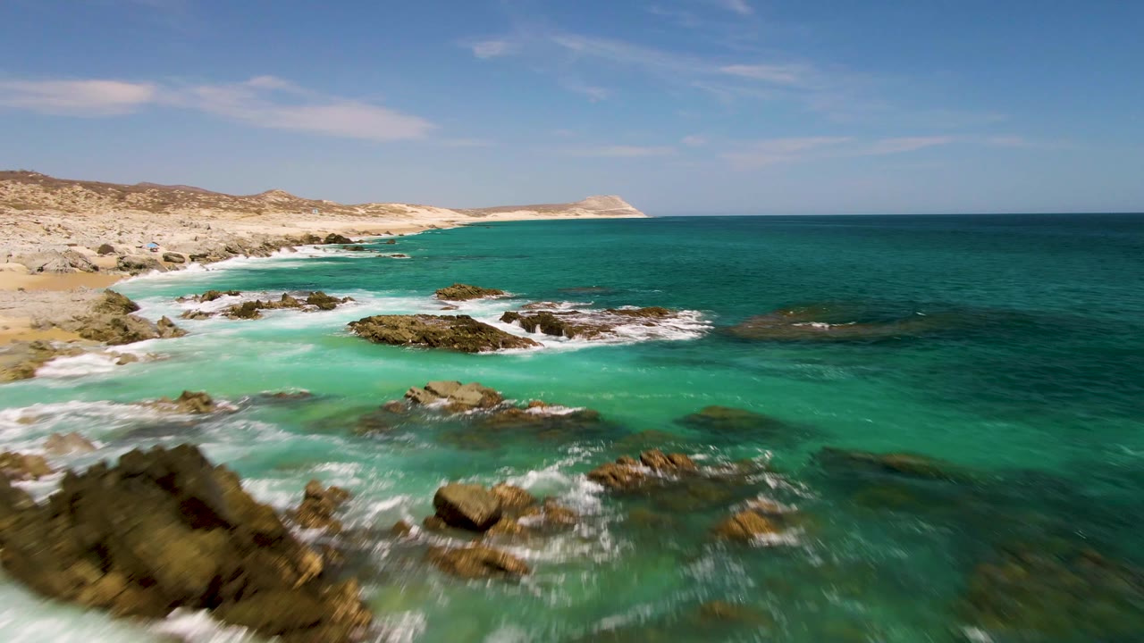 Aerial frame at Buzzards Beach showing white water wrapping around several exposed rocks, a sandy shoreline to the left, distant hills, and no visible people.