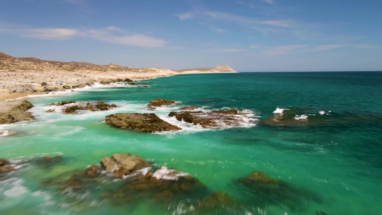 Aerial view of Buzzards Beach with a wider stretch of turquoise ocean, scattered rock outcrops, pale beach, low arid hills, and no visible people.