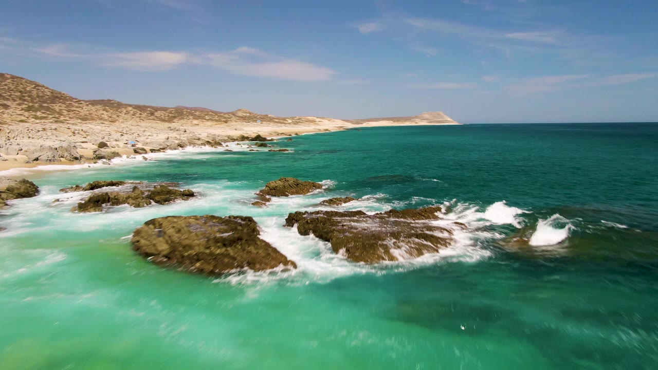 Aerial frame from Buzzards Beach showing white surf hitting dark rocks in the foreground, green-blue ocean across the midground, sandy coast to the left, and no visible people.