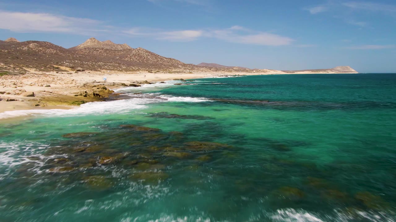 Aerial frame at Buzzards Beach, Baja California Sur, showing turquoise shallows over dark rocks, a sandy beach, distant mountains, blue sky, and no visible people.