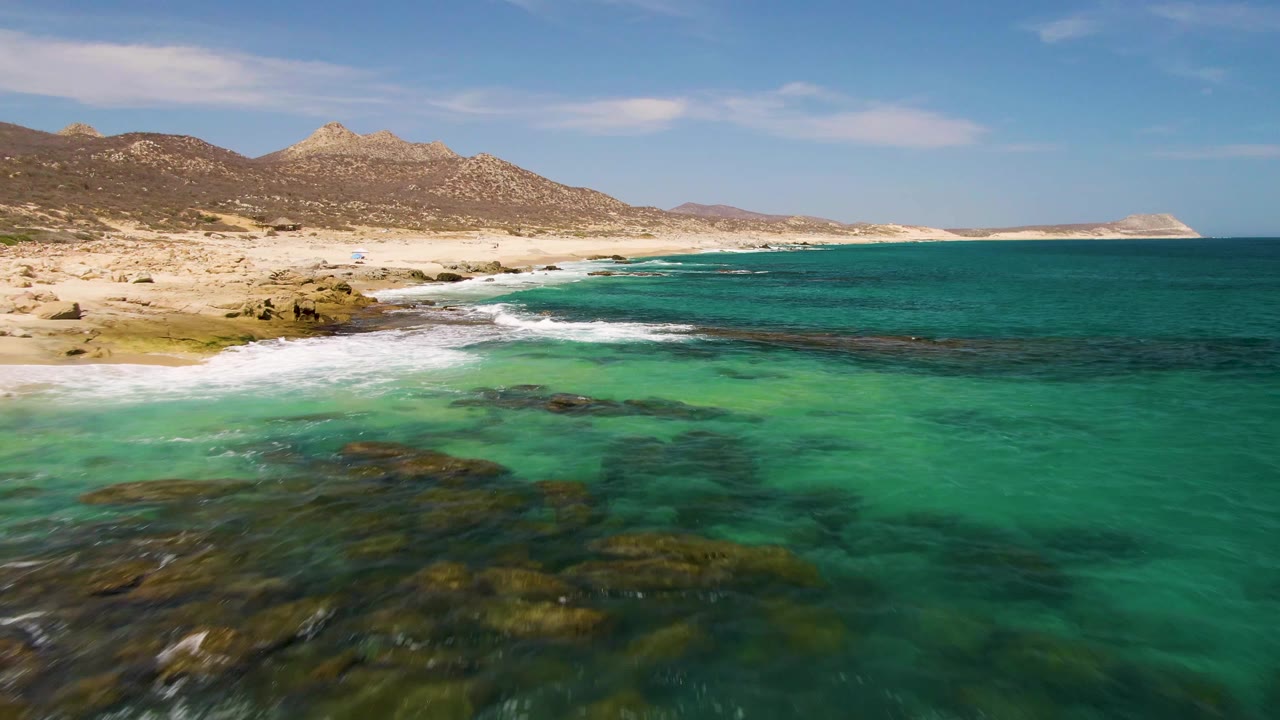 At 0:01, the Buzzards Beach shoreline remains in a high aerial view with dark underwater rocks centered in green-turquoise water, surf along the sand, mountains, and no visible people.
