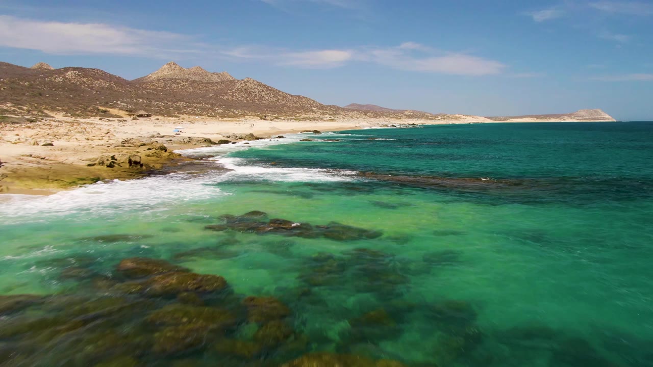 At 0:02, clear water spreads over larger dark rock patches in the foreground while white surf lines the sandy Buzzards Beach shore below brown mountains, with no visible people.