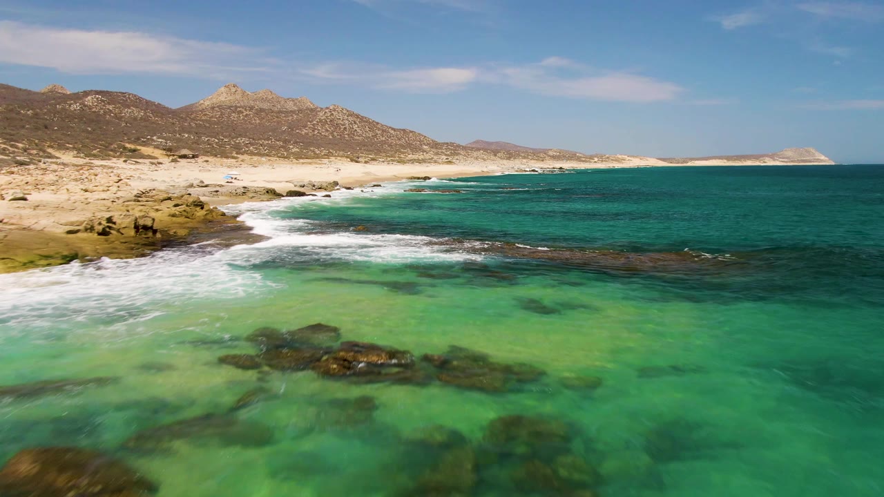 White waves roll toward a sandy beach with turquoise shallows and mountains behind.