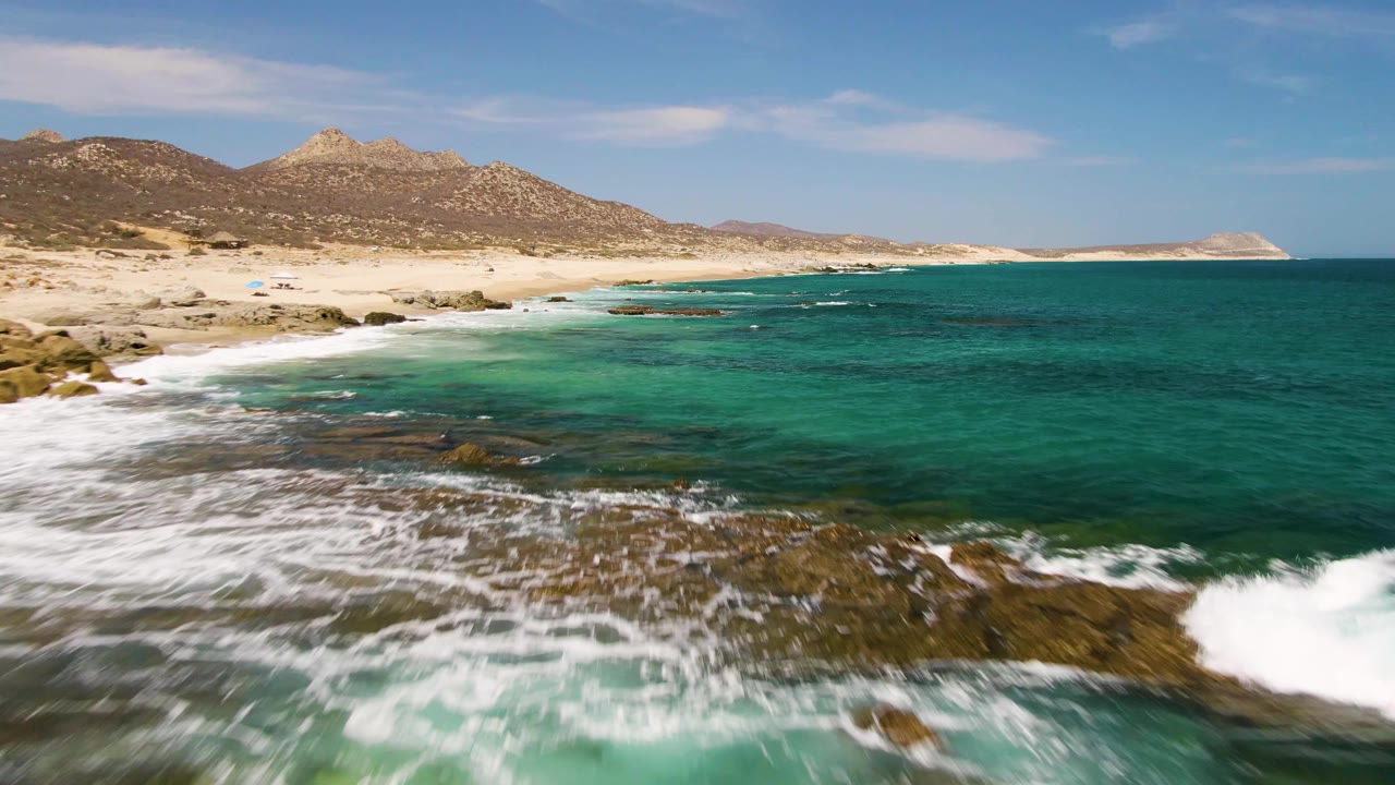 Aerial view at Buzzards Beach, Baja California Sur, with white waves breaking over dark rocks in the foreground, turquoise water, pale sand, low mountains, and no visible people.