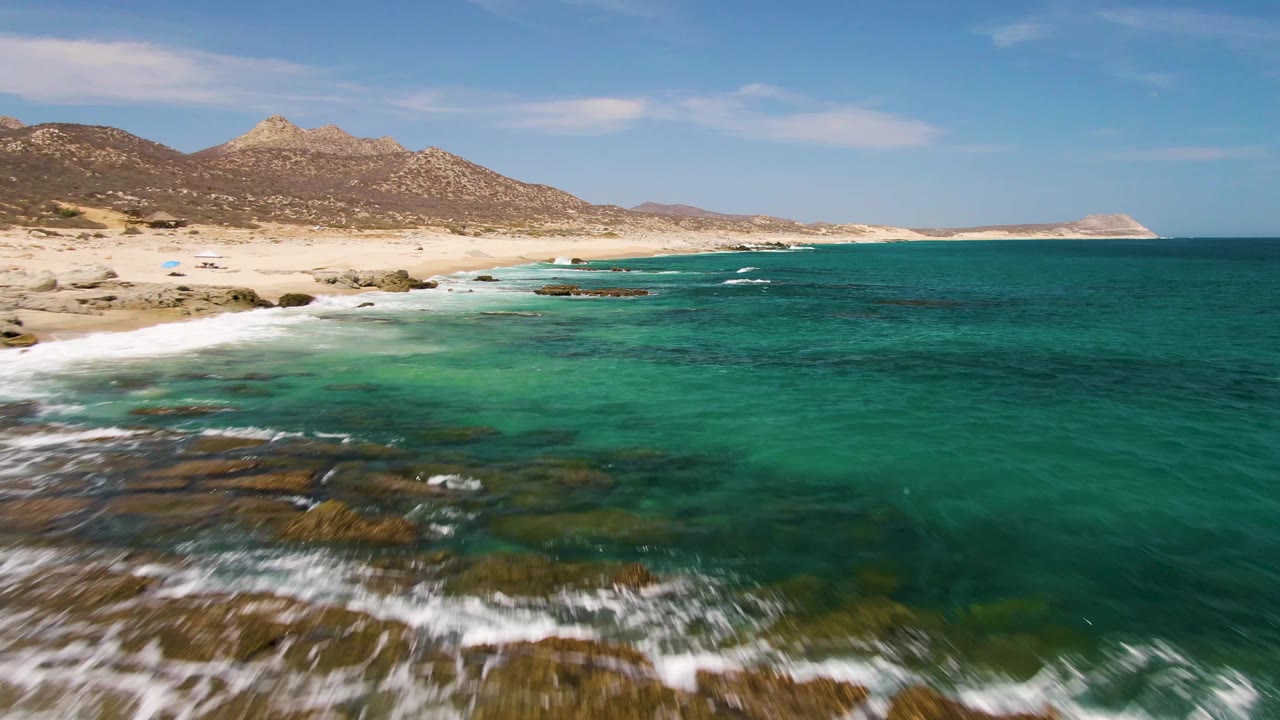 Aerial frame over Buzzards Beach showing clear green-turquoise shallows across submerged rocks, small wave lines near the sand, distant mountains, and no visible people.