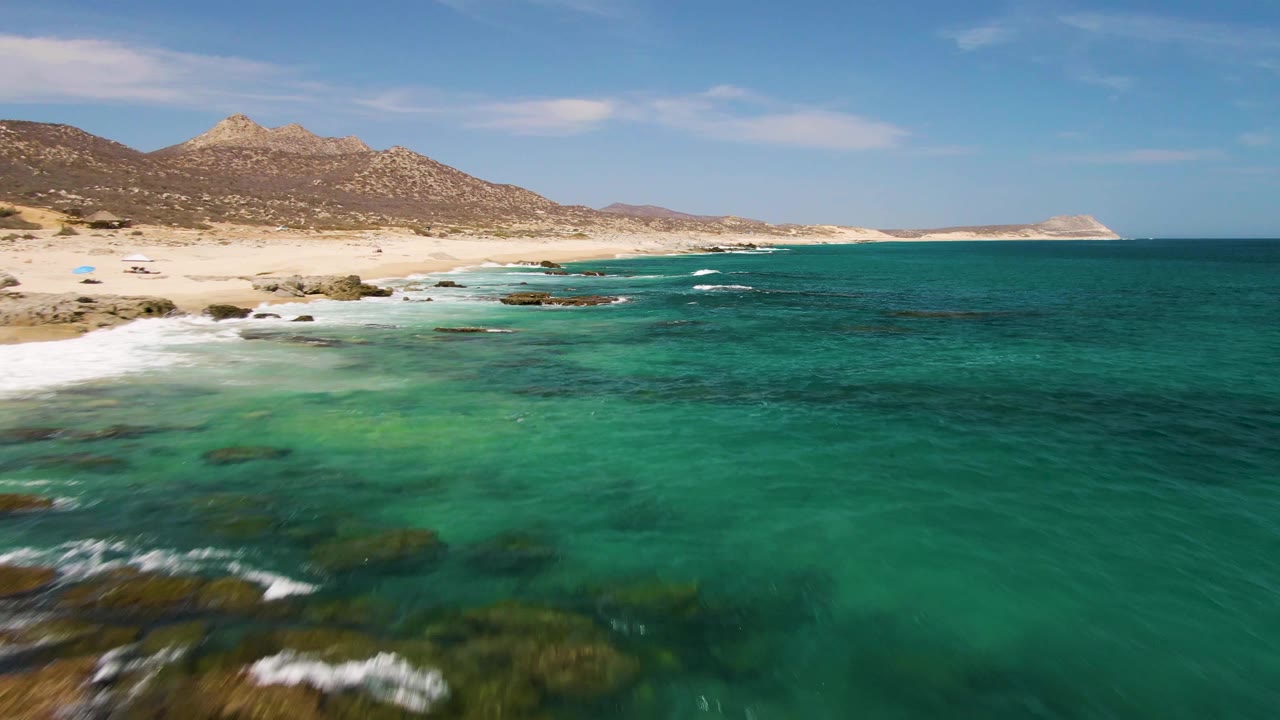 Aerial coastal view at Buzzards Beach with more open turquoise water in the foreground, scattered submerged rocks near shore, pale sand, low mountains, and no visible people.