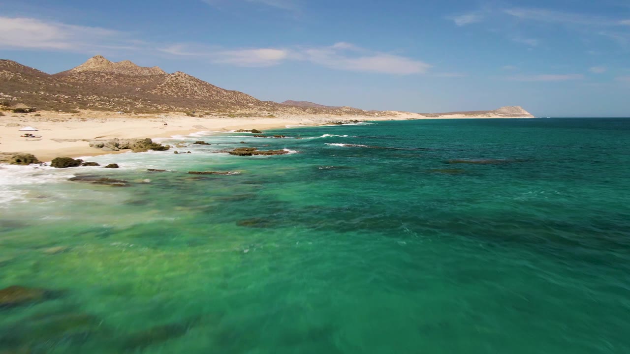 Shallow green water leads to a sandy beach with rocks and mountains.