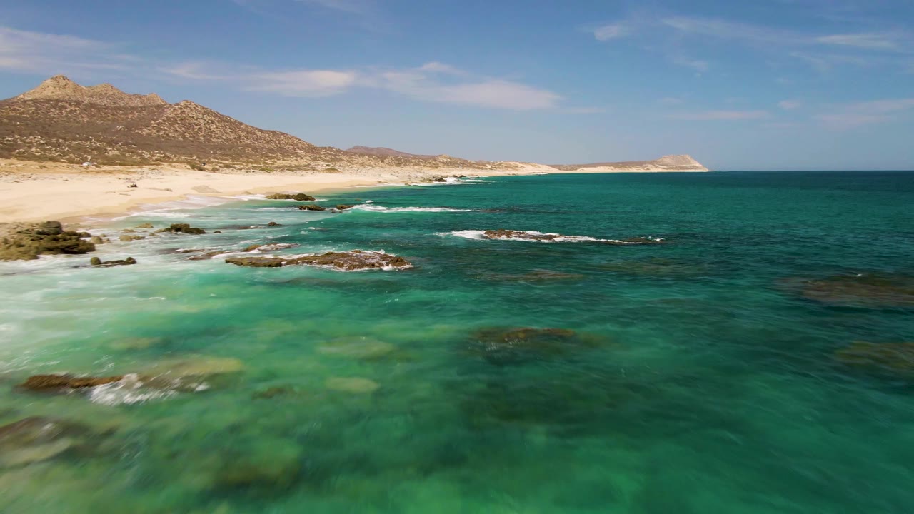 Elevated view of Buzzards Beach with turquoise shallows over submerged rocks, a sandy beach at left, mountains in the background, and no visible people.