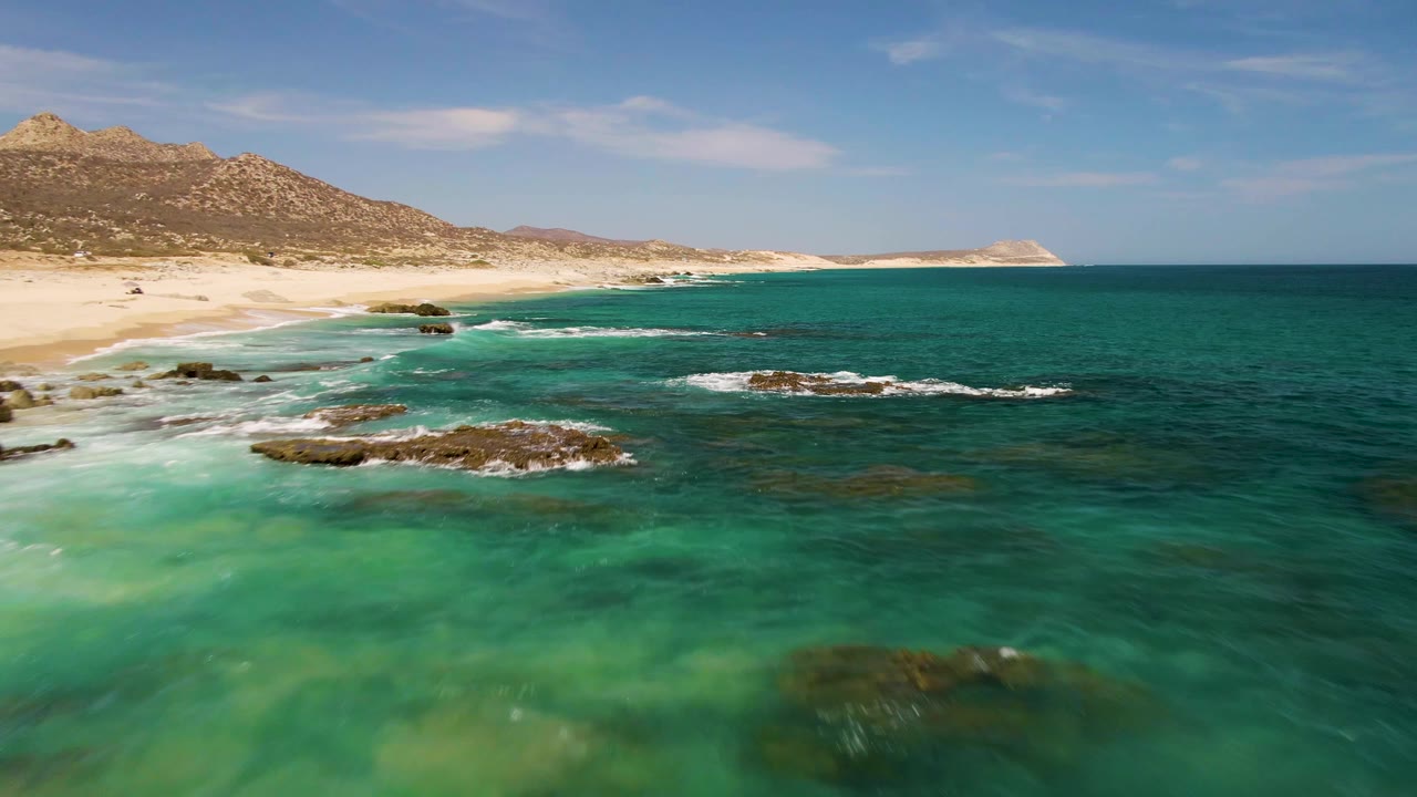 Buzzards Beach shoreline with green-blue water spreading over shallow rocks, small breaking waves near the sand, distant mountains, and no visible people.