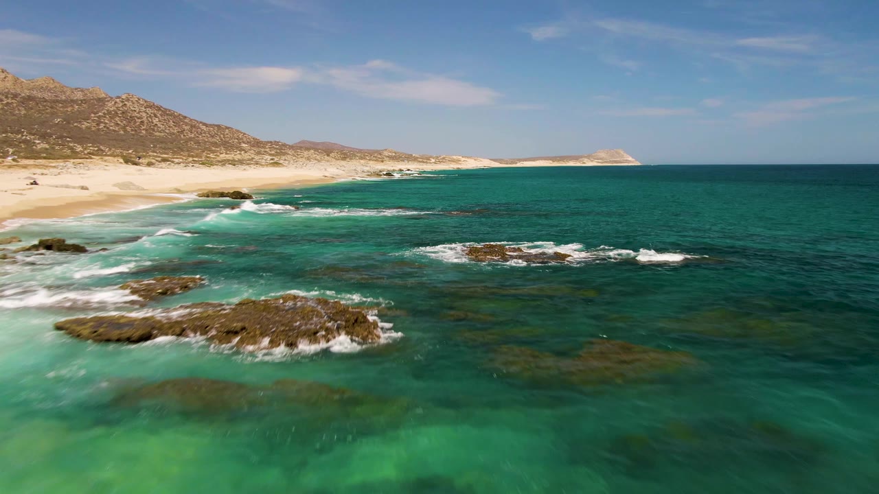 Aerial coastal frame at Buzzards Beach with exposed rocks nearer the center, whitewater curling around them, pale sand to the left, and no visible people.