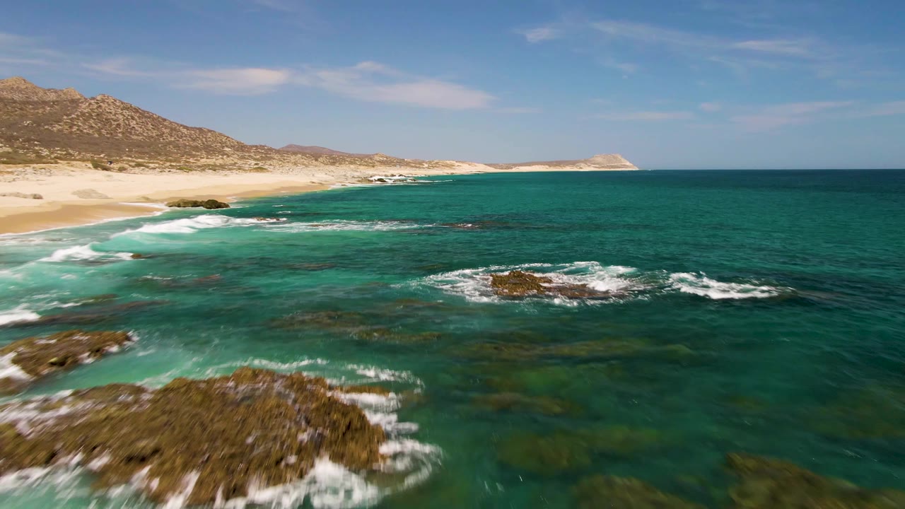 Buzzards Beach view with foreground rocks and white foam at the lower edge, clear turquoise water across the middle, sandy beach at left, and no visible people.