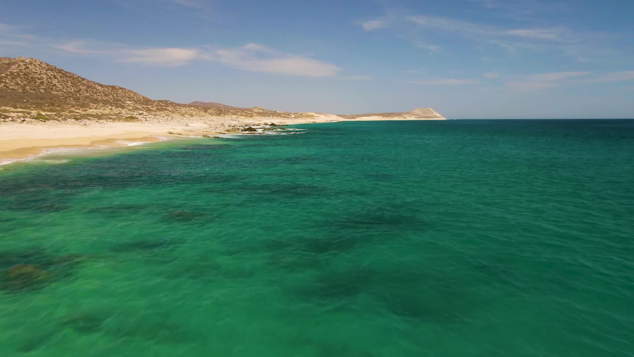 Green-blue ocean water with darker seabed patches leads toward an empty sandy beach and rocky hills at Buzzards Beach, with no visible people.