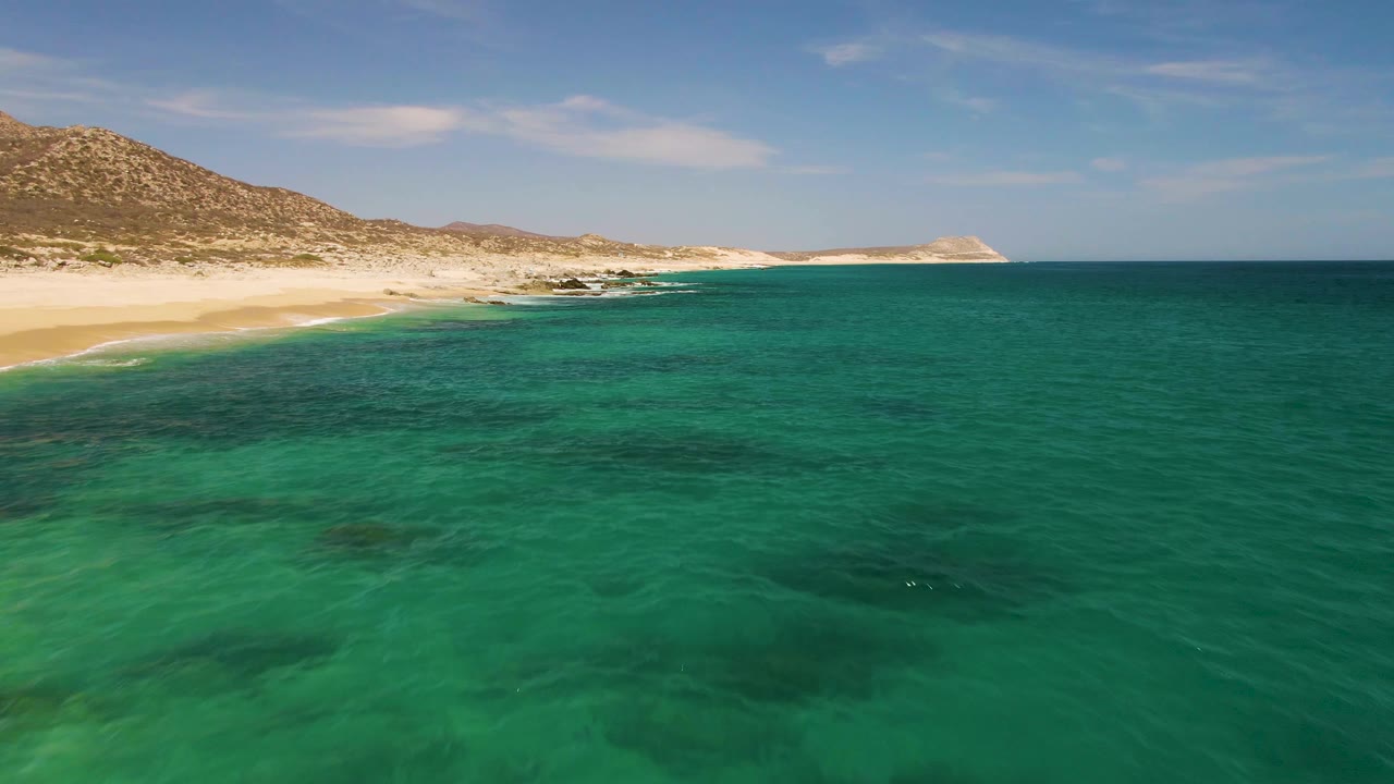 Small waves meet the sandy shoreline at Buzzards Beach as turquoise water and dark underwater rock patches fill the lower frame, with no visible people.