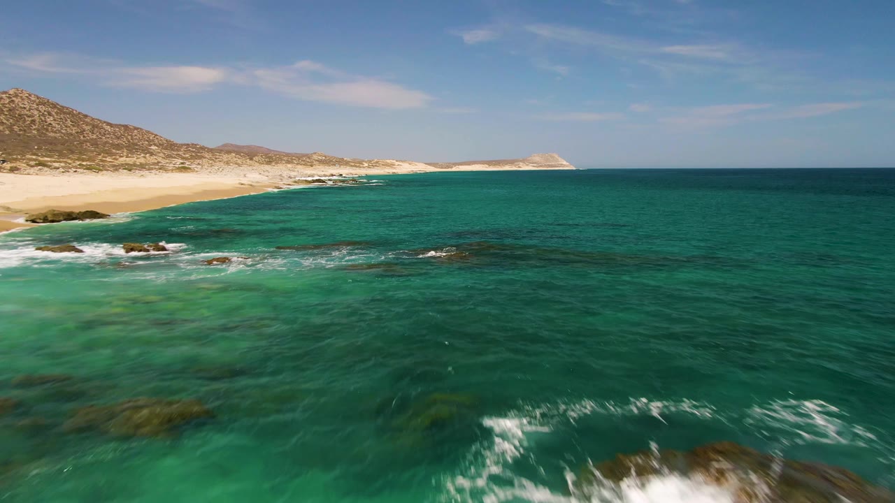 Elevated view over Buzzards Beach in Baja California Sur with turquoise water, submerged rocks, a sandy shoreline to the left, low hills in the distance, and no visible people.