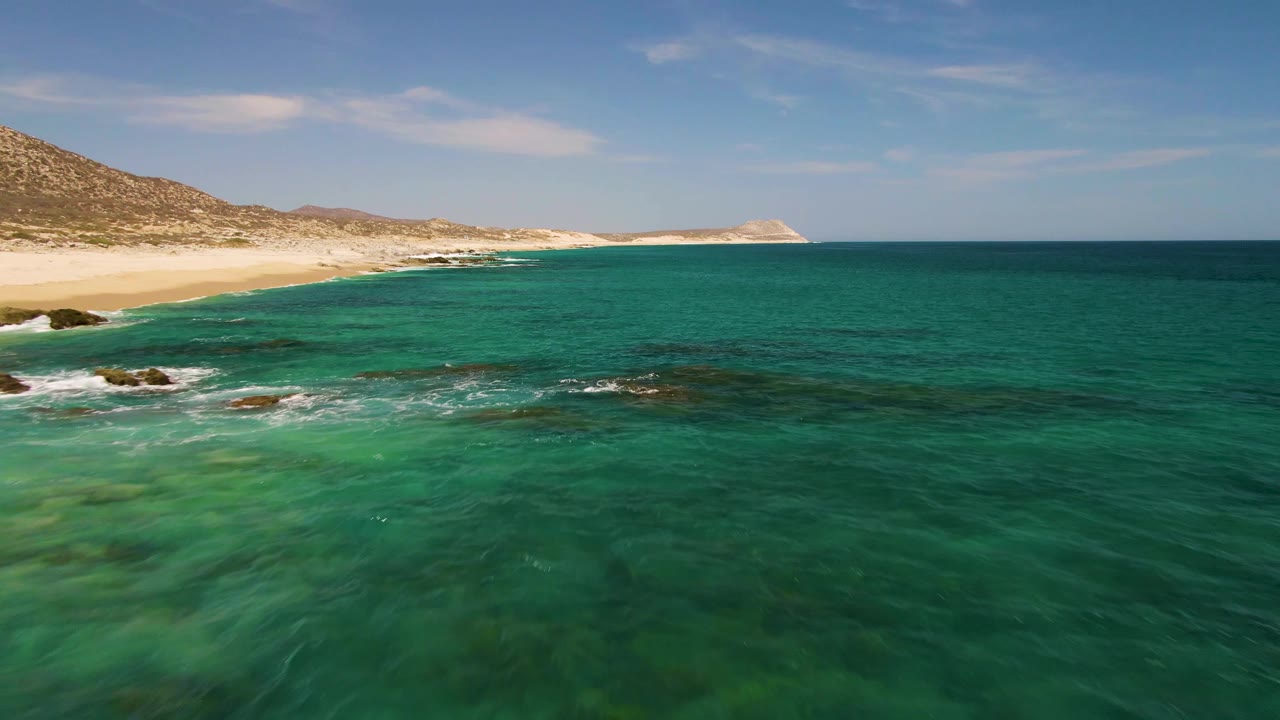 Clear green-blue water with shoreline, rocks, sand, and distant hills.