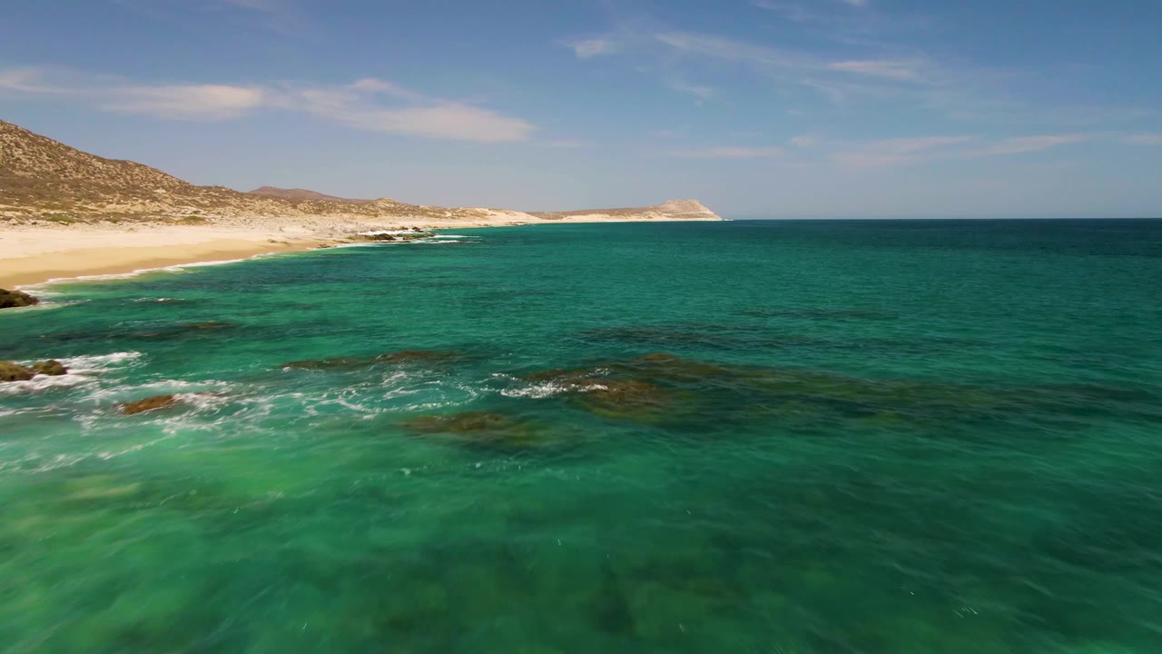 Aerial-looking frame at Buzzards Beach with turquoise ocean in the foreground, small white breaks over rocks near shore, pale sand to the left, low hills beyond, and no visible people.