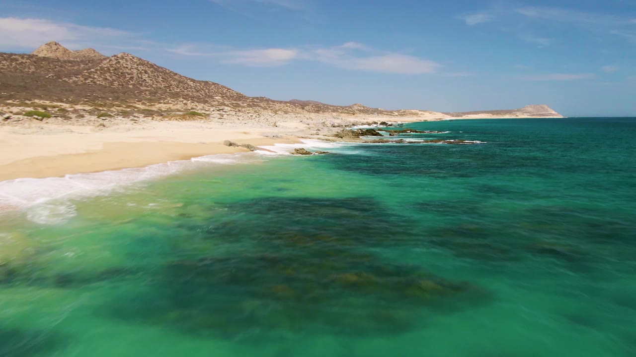 Buzzards Beach shoreline seen from above, with a stronger band of white surf at the left edge, green-blue shallows in the foreground, low rocky hills beyond the sand, and no visible people.