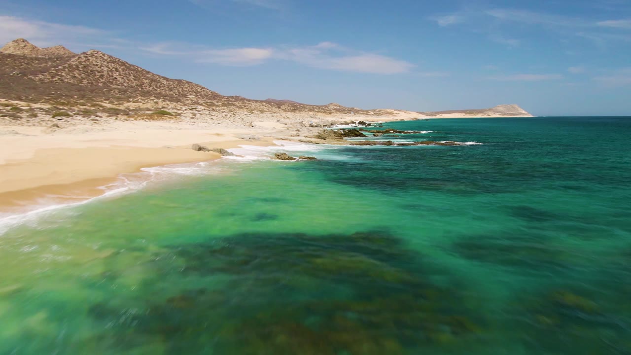 Mid-clip frame at Buzzards Beach with turquoise water filling most of the view, pale beach along the left, scattered dark submerged areas under the surface, rocky hills behind the sand, and no visible people.