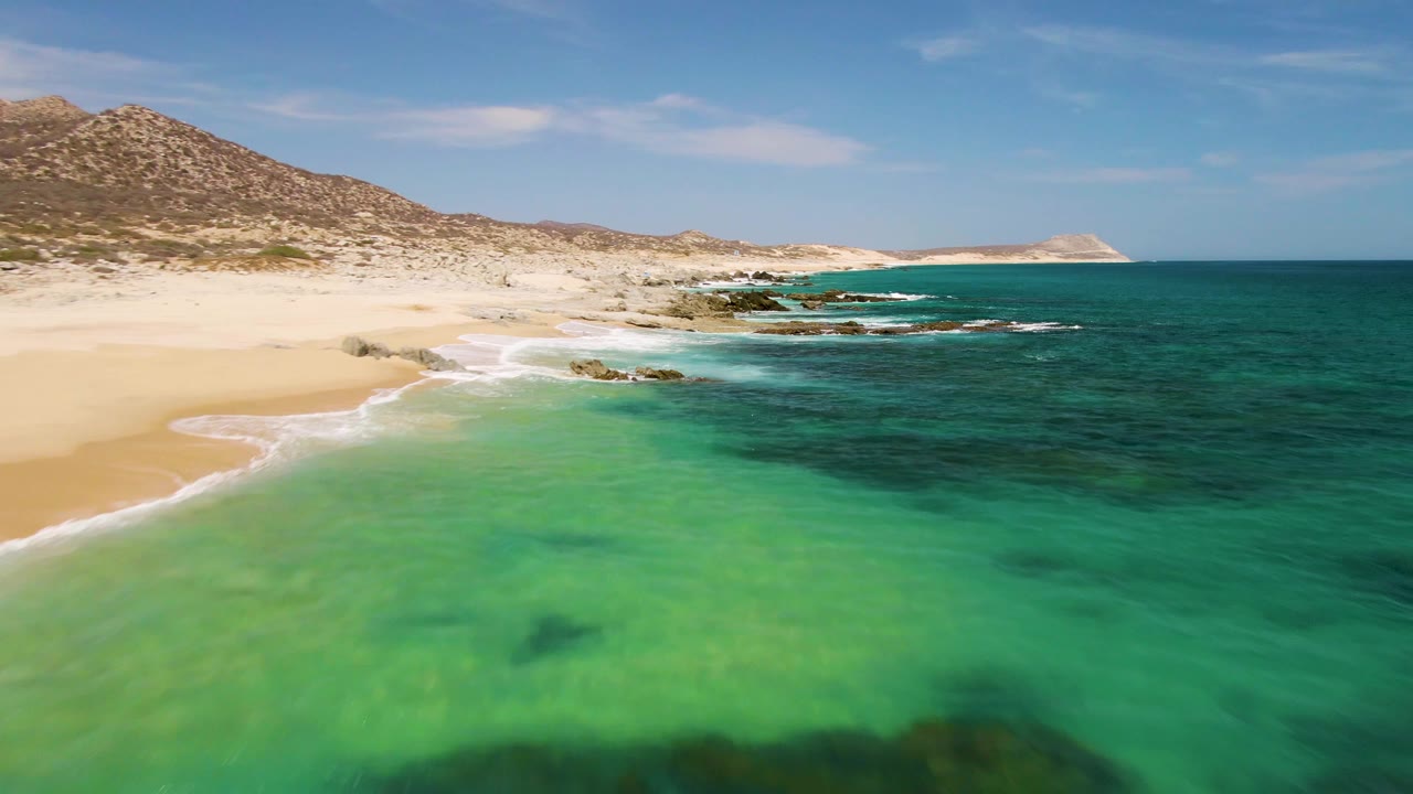 Buzzards Beach coast with the sandy shore sweeping from the lower left, clear green water in the foreground, darker blue-green ocean to the right, rocky points along the midground, and no visible people.