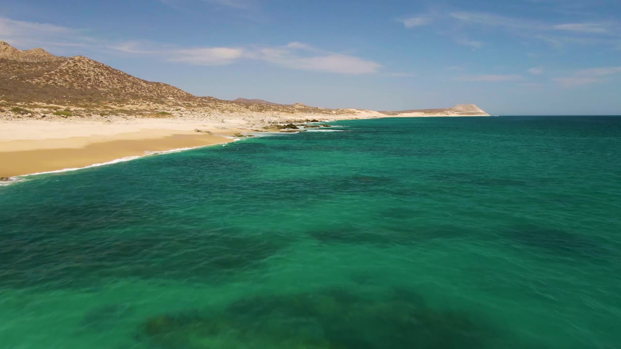 At Buzzards Beach in Baja California Sur, turquoise ocean fills the foreground while a pale sandy shore and rocky hills stretch across the background with no visible people.