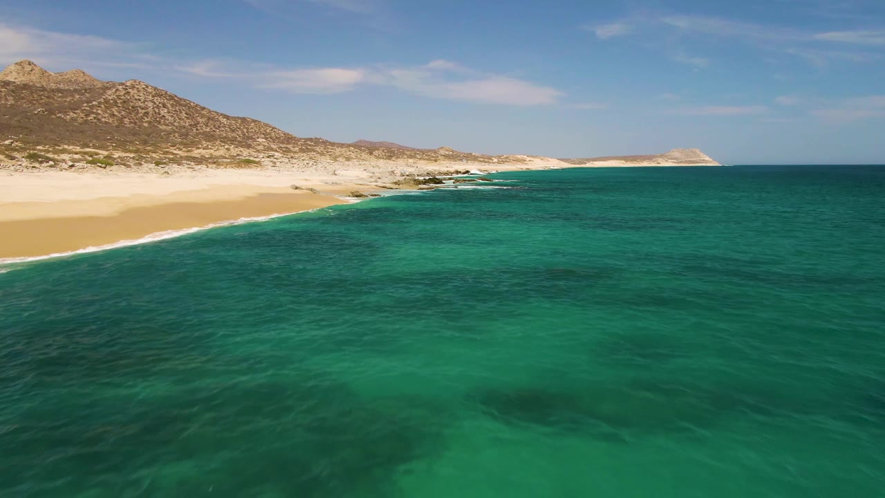 One second into the clip at Buzzards Beach, green-blue water covers most of the frame as small white surf lines reach the sand below rocky Baja California Sur hills with no visible people.