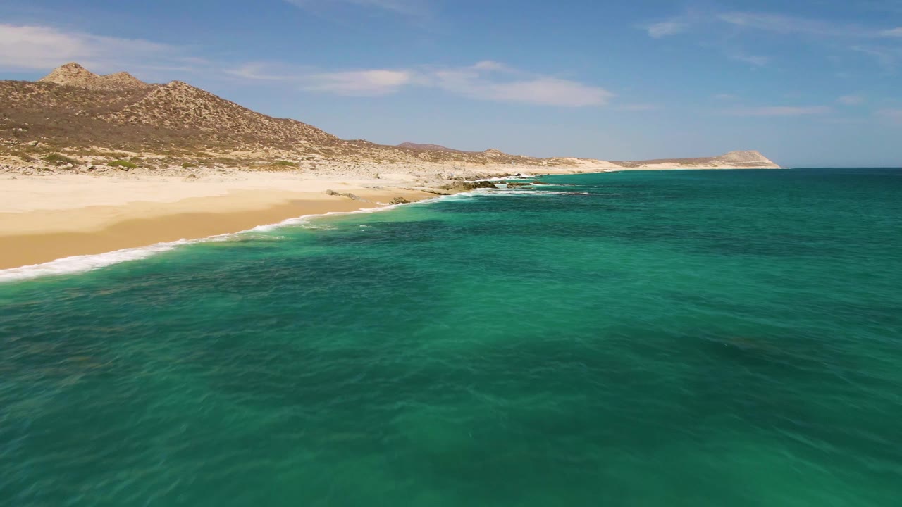 Sandy beach curves beside turquoise water with rocky coastal mountains behind.