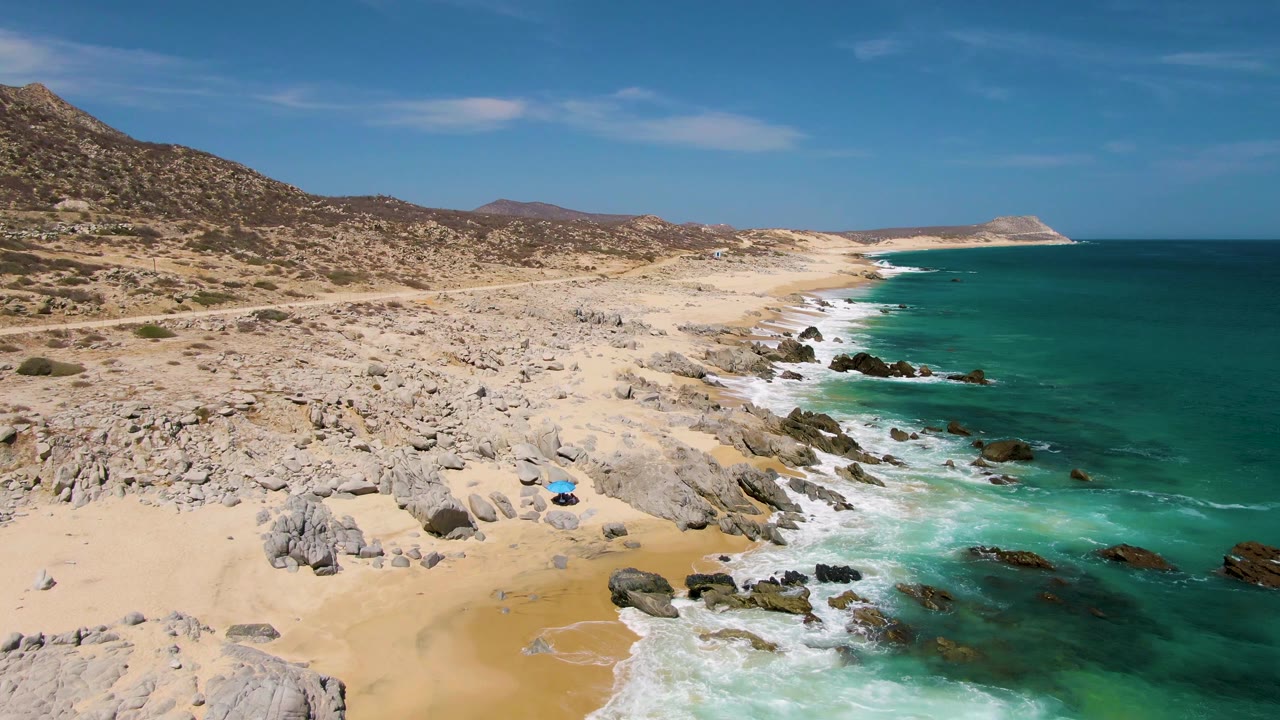 Aerial view of sandy beach, rocks, turquoise ocean, and dry hills.