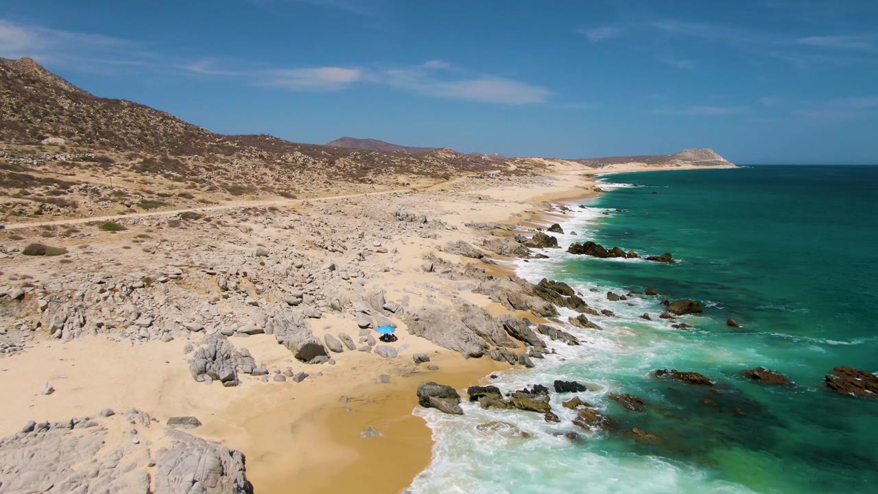 Buzzards Beach shoreline seen from above with scattered rocks on sand, white surf curling into turquoise water, dry hills inland, and no visible people.