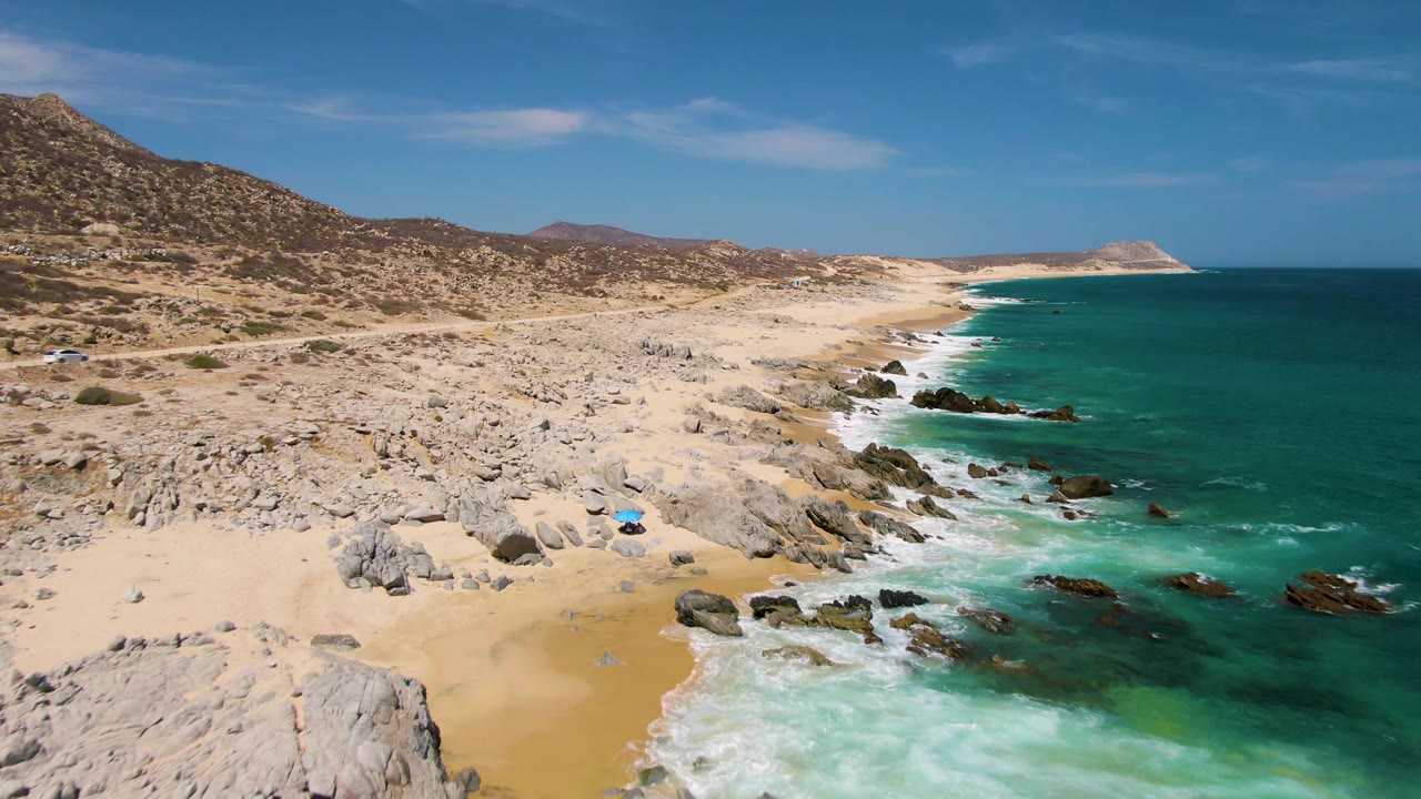 High oblique view of Buzzards Beach with a larger sweep of turquoise ocean on the right, pale rocky sand on the left, distant headland, and no visible people.