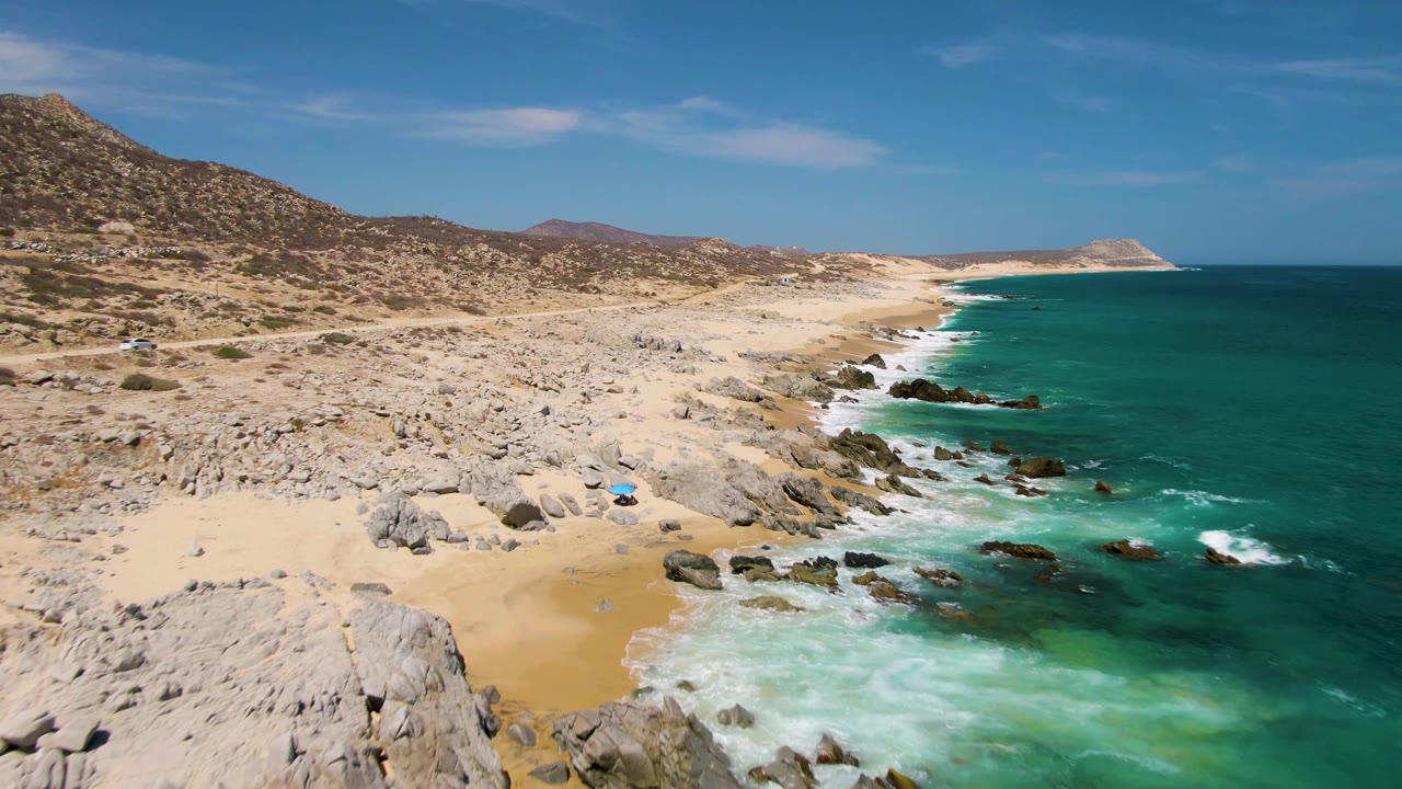 Buzzards Beach appears from a slightly closer aerial angle with large foreground rocks, sand pockets, foamy turquoise surf, inland hills, and no visible people.