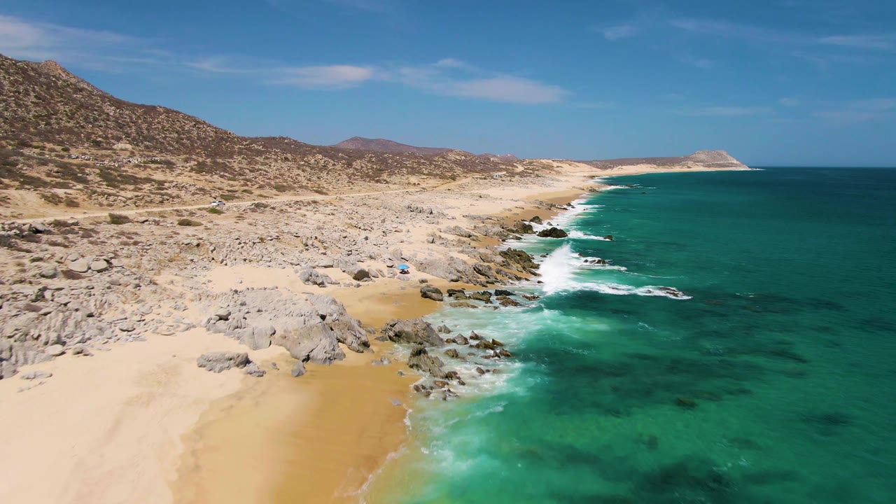 Aerial view along Buzzards Beach with the shoreline curving into the distance, white waves breaking on rocks, dry hills inland, and no visible people.