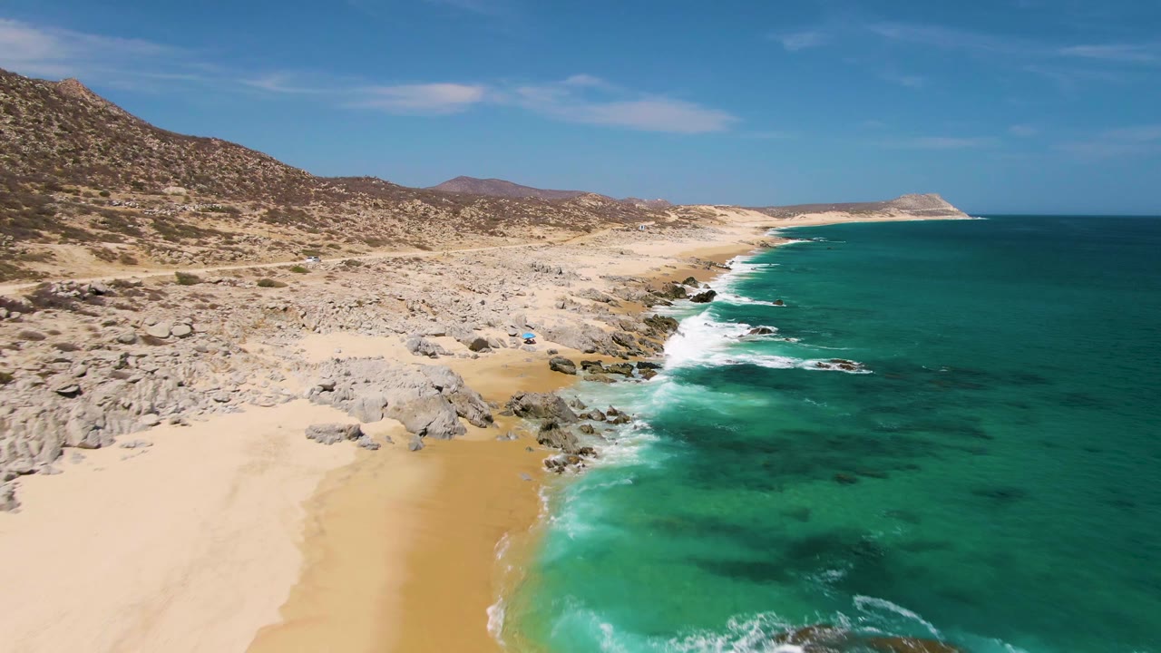 Buzzards Beach aerial frame showing a sandy cove in the foreground, green-blue water to the right, scattered shoreline boulders, distant hills, and no visible people.